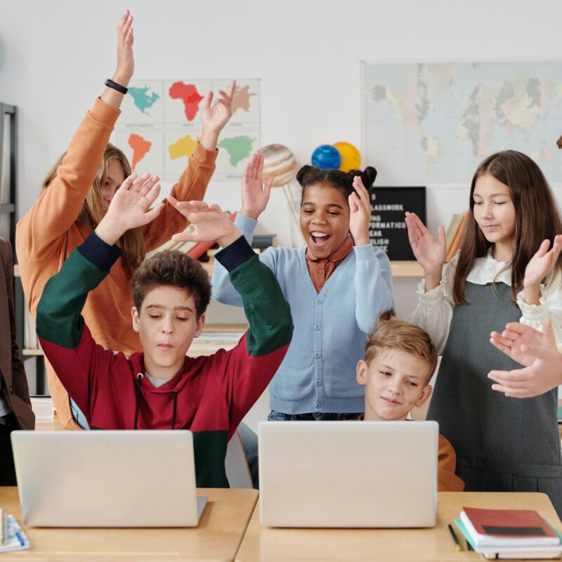 A group of cheerful students and their teacher celebrate a successful classroom activity while gathered around laptops.