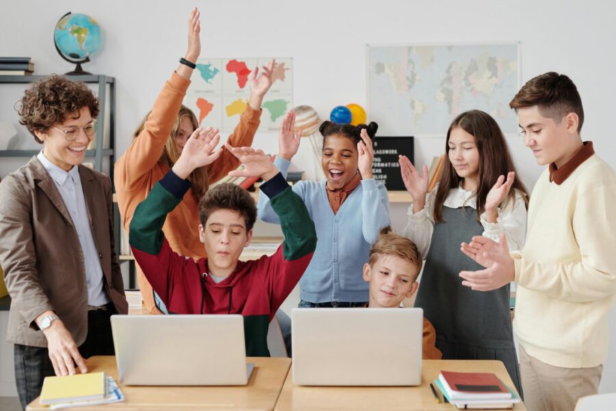 A group of cheerful students and their teacher celebrate a successful classroom activity while gathered around laptops.
