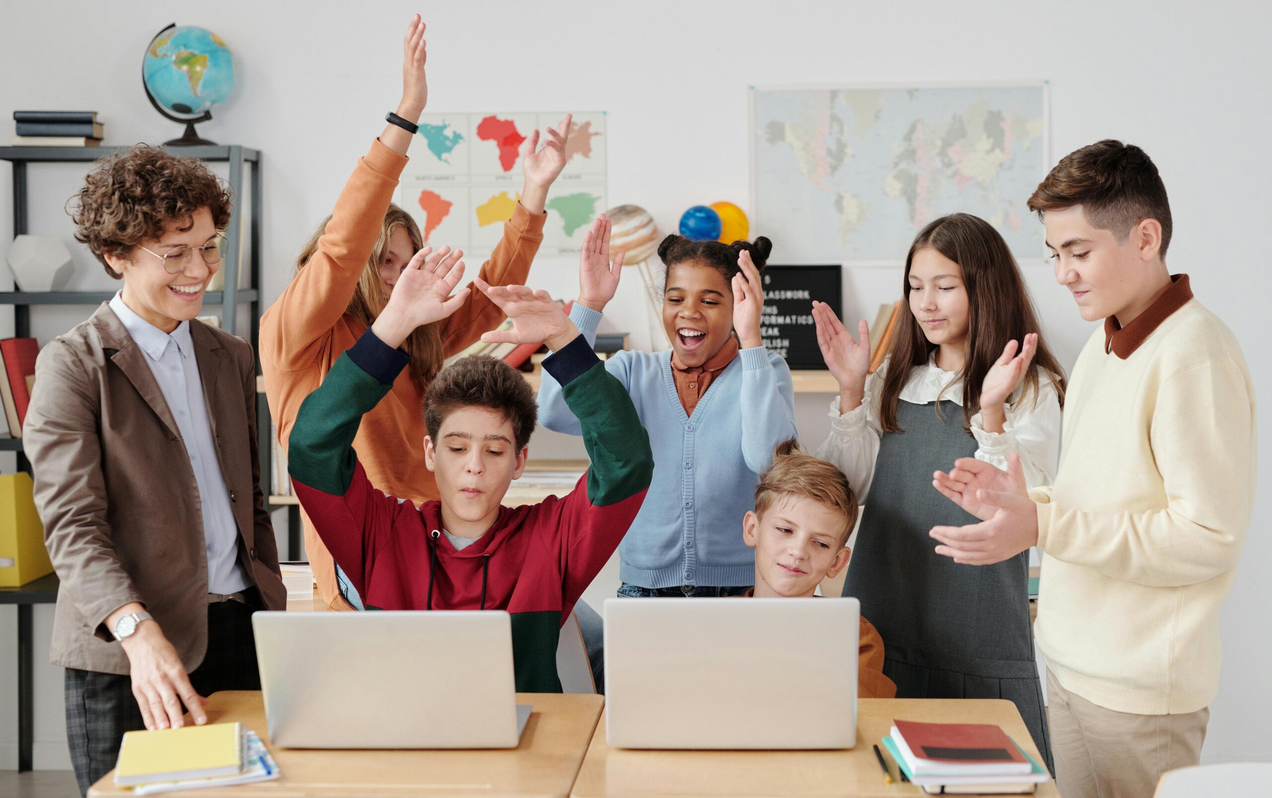 A group of cheerful students and their teacher celebrate a successful classroom activity while gathered around laptops.