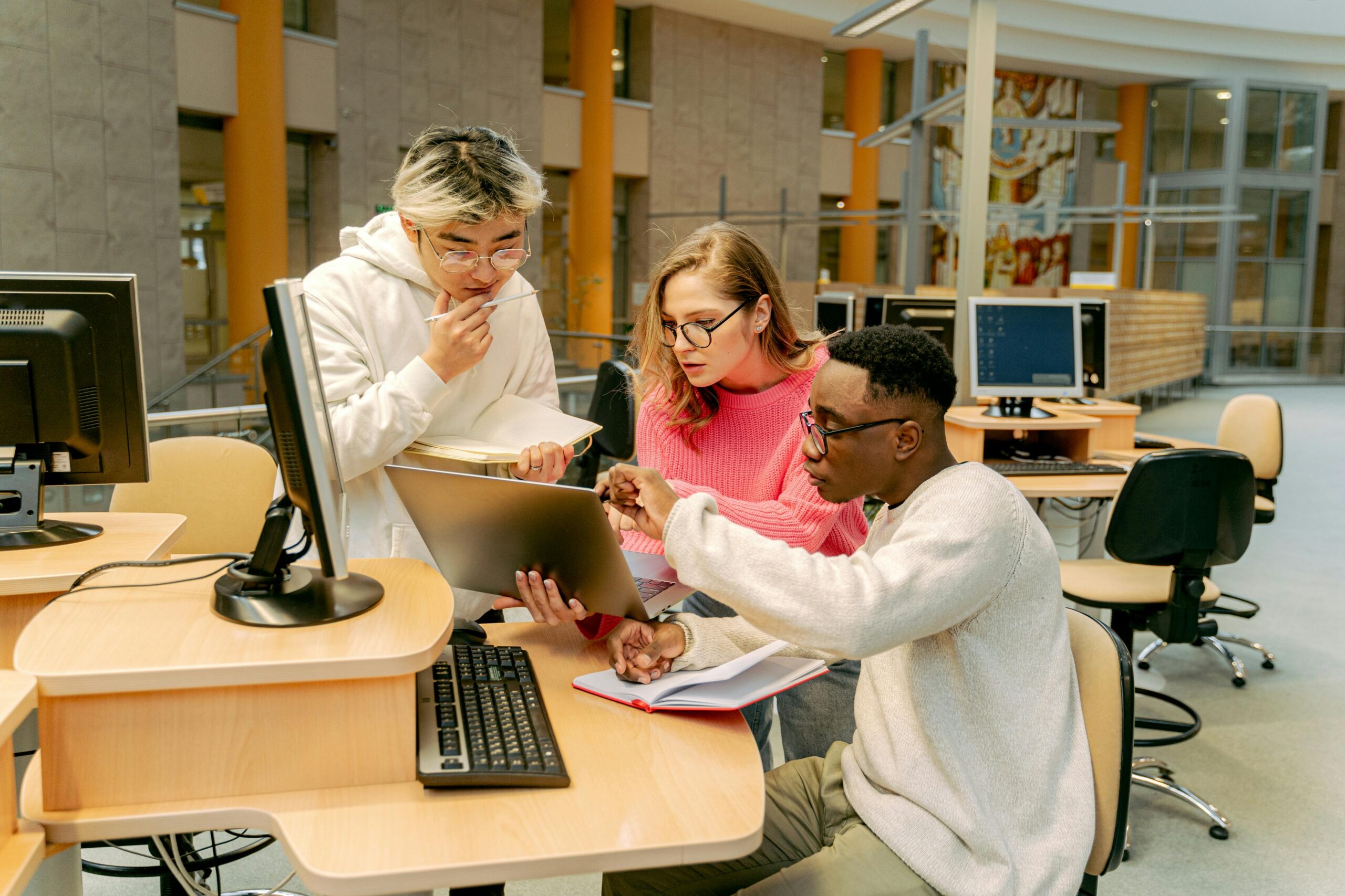 Three focused students collaborate on a project using a laptop and computer in a computer lab setting.