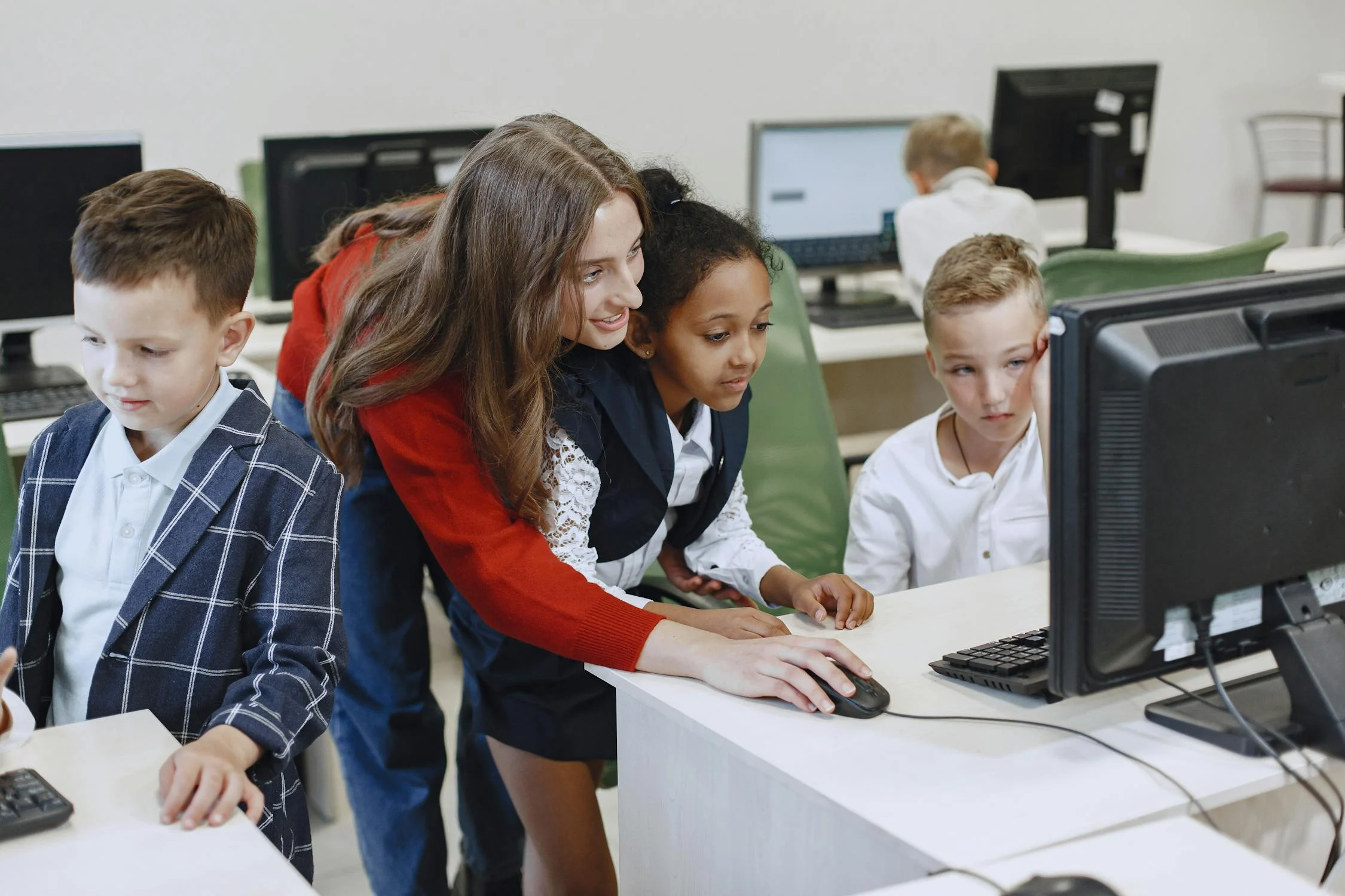 A teacher helps a group of young students work on desktop computers in a classroom.