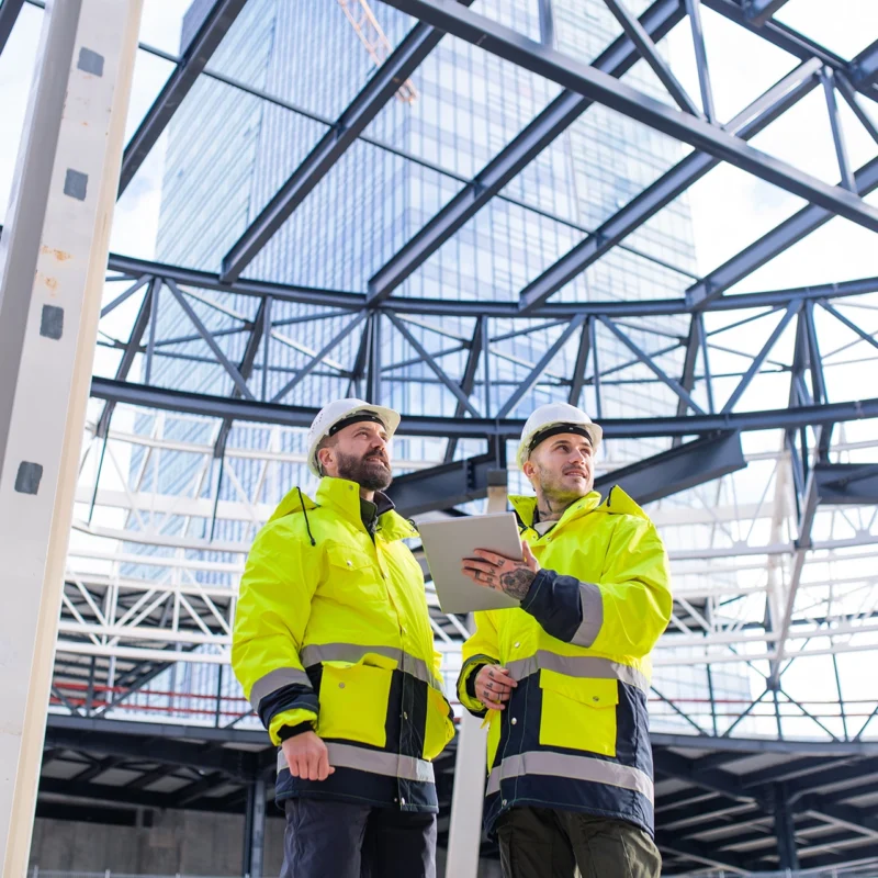 Two construction professionals in high-visibility gear review project data on a tablet at a commercial jobsite, highlighting the use of business process automation tools in the construction industry.