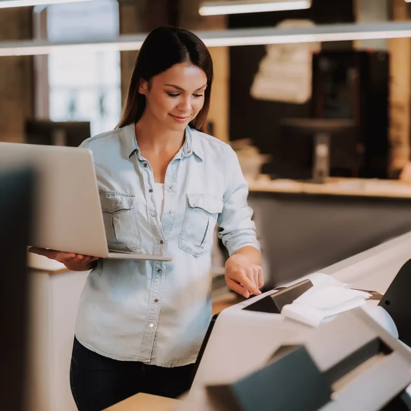 Woman using a multifunction printer while holding a laptop in a modern office, representing unified technology solutions, document workflow automation, and managed IT services.