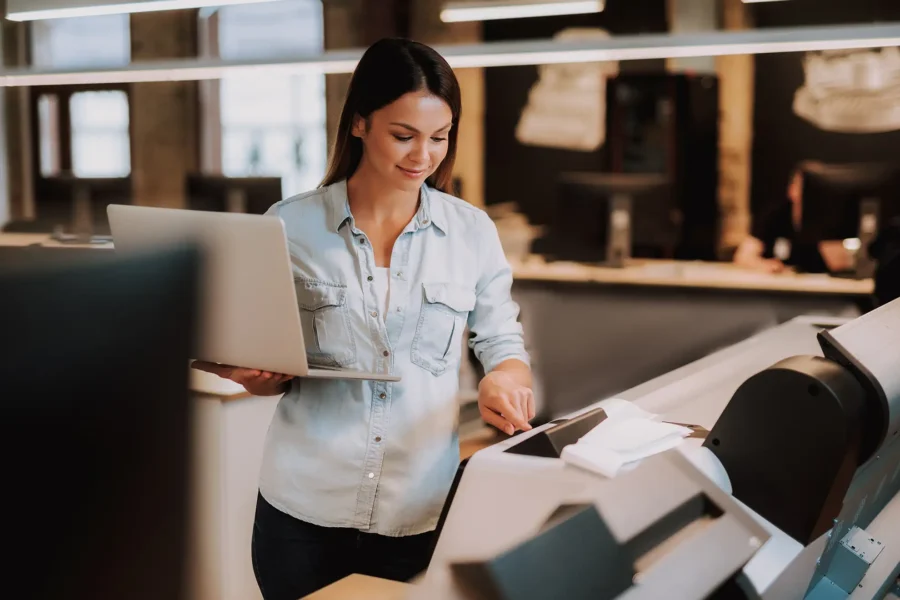 Woman using a multifunction printer while holding a laptop in a modern office, representing unified technology solutions, document workflow automation, and managed IT services.